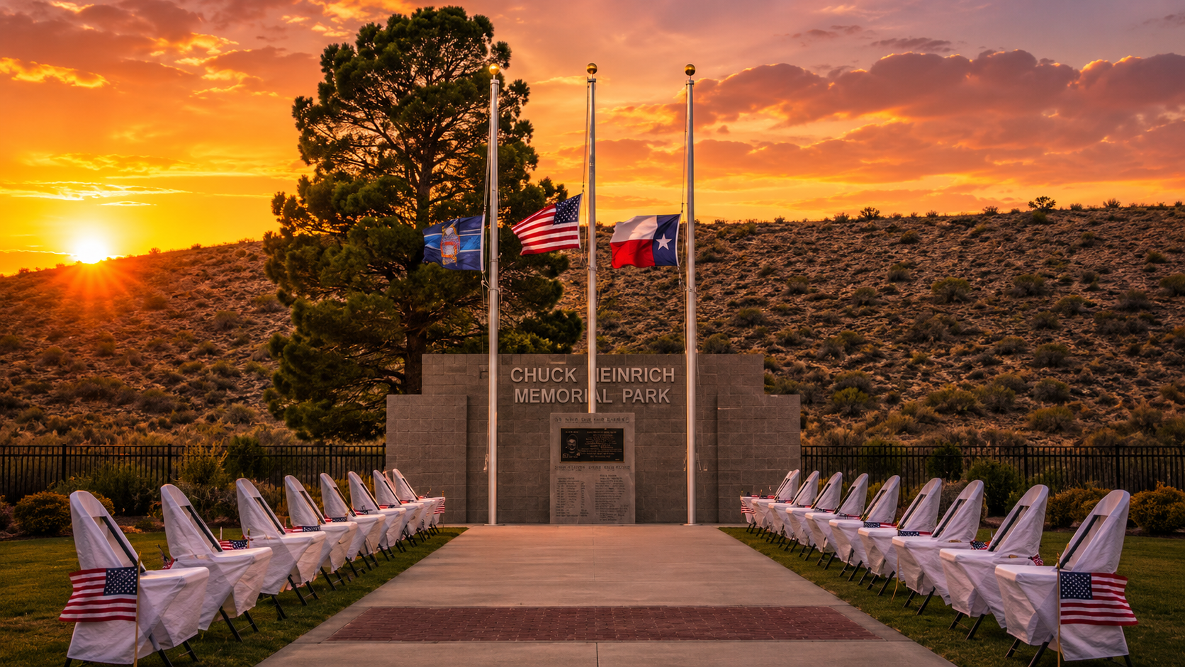 A memorial monument at Chuck Heinrich Memorial Park at sunset, flanked by flags at half-mast and rows of chairs draped in white